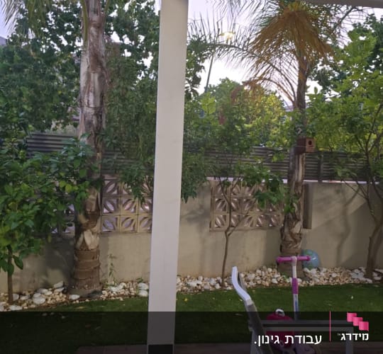 עצים ושיחים בגינה עם דשא מלאכותי ואבנים לבנות